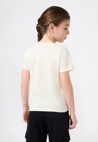 Jauna mergaitė su pintinėmis plaukais dėvi paprastą kreminės spalvos marškinėlį su trumpomis rankovėmis ir juodas kelnes, stovinti šonu su galva pasukta žemyn.