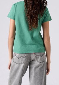 Personne aux longs cheveux bouclés portant un t-shirt vert à manches courtes et un jean gris clair, debout de dos sur un fond uni.