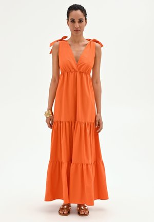 Femme portant une robe maxi orange sans manches avec bretelles à nouer et jupe à volants, accessoirisée de bracelets dorés et de sandales plates.