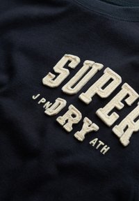 Suéter azul marino con letras bordadas texturizadas de color crema que dicen "SUPER DRY ATH". Tejido suave con escote redondo.