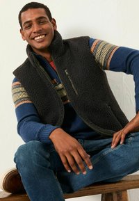 Schwarze Fleece-Weste, die über einem blauen Pullover mit gestreiften Akzenten getragen wird. Das Modell sitzt, trägt eine blaue Jeans und braune Schuhe und lächelt vor einem neutralen Hintergrund.