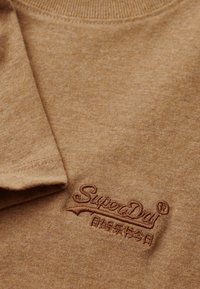 Felpa corta in cotone marrone, con maniche corte, caratterizzata da un colletto a coste e un logo ricamato "SuperDry" in marrone scuro. Texture liscia.