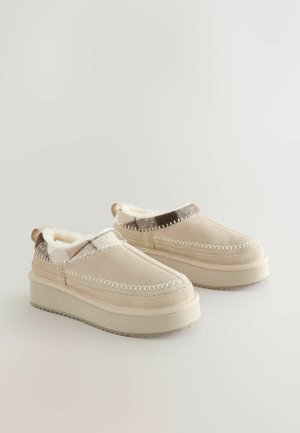 Beige Slip-On-Schuhe mit einem weichen, kuscheligen Futter, ausgestattet mit einer leicht erhöhten Plattform und dekorativen Nähten entlang der Ränder.