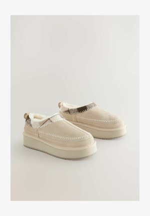 Beige Slip-On-Schuhe mit einem weichen, kuscheligen Futter, ausgestattet mit einer leicht erhöhten Plattform und dekorativen Nähten entlang der Ränder.