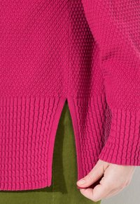 Mano che aggiusta la fessura laterale di un maglione a maglia testurizzata rosa sopra dei pantaloni verde oliva.
