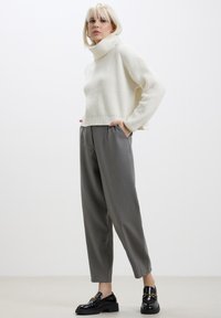 Pull à col roulé blanc à côtes associé à un pantalon gris fuselé et des mocassins noirs. Le pull a un design cropped ; les chaussures présentent une touche dorée.