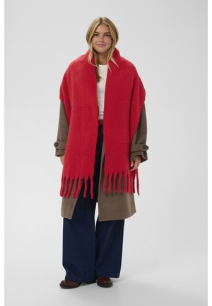 Red wool scarf with fringed edges draped over a brown coat, paired with dark blue wide-leg jeans and brown shoes. Plain background.