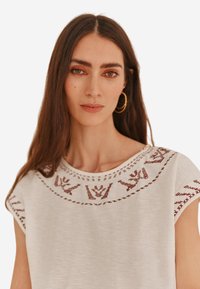 Haut blanc à manches courtes avec un motif brodé rouge autour de l'encolure. Texture douce, col rond et légère drapé.
