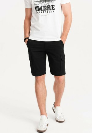 Schwarze Cargo-Shorts mit Seitentaschen, aus robustem Stoff gefertigt. Getragen mit einem weißen T-Shirt und lässigen weißen Sneakers.