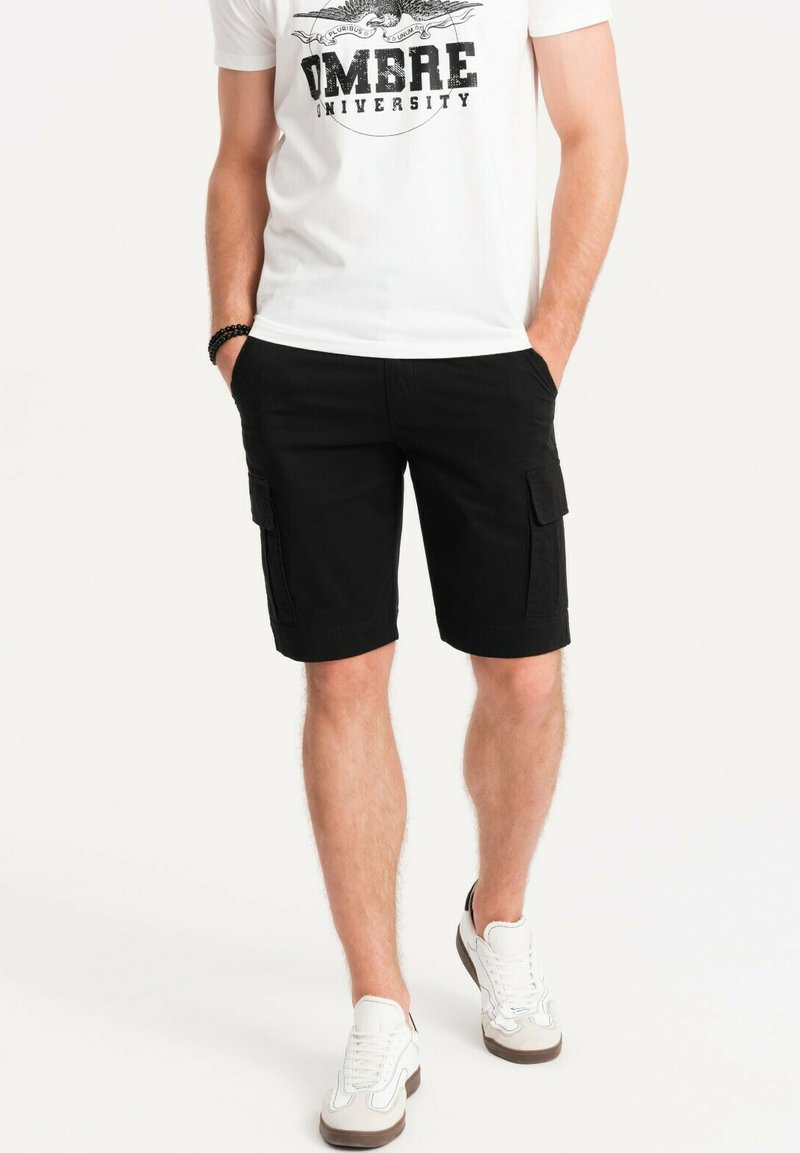 Pantaloni cargo neri con tasche laterali, realizzati in tessuto resistente. Indossati con una t-shirt bianca e sneakers bianche casual.