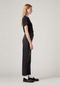 Black T-shirt, high-waisted black jeans, white socks, black loafers. Smooth fabric, straight leg design, minimal detailing. Side profile view.