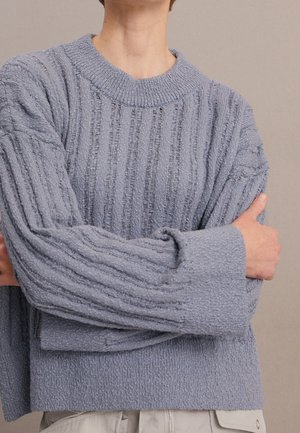 Pull en tricot bleu clair avec texture côtelée, poignets larges et encolure arrondie, offrant une coupe décontractée et un matériau doux.