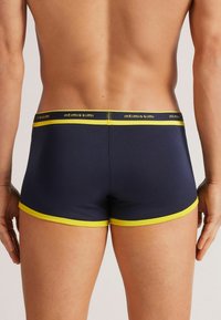 Mörkblå herrboxershorts med gul midjeband och detaljer, tillverkade av ett slätt tyg, med logodetaljer på midjebandet.