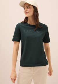 Camiseta de algodón verde oscuro con mangas cortas, que presenta el texto en relieve "CIELOS ETERNOS" en el frente. Combinada con pantalones beige claros.