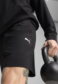 Shorts sportive negre, confectionate dintr-un material neted, cu un logo alb Puma pe partea din stânga jos. O mână ține o kettlebell neagră.