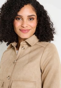 Veste en daim beige avec un col, deux poches avant et des boutons. Texture lisse et coupe ajustée visibles.
