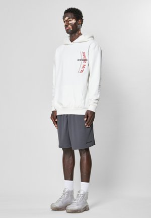 Hvid hættetrøje med frontlomme og rød tekstdetalje, parret med grå mesh-shorts og grå snøre støvler. Ensfarvet baggrund.