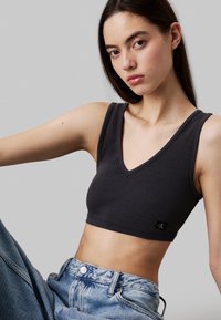 Top corto negro acanalado con diseño de cuello en forma de V, que presenta un pequeño logo en el lateral, combinado con jeans de mezclilla azul de tiro alto.