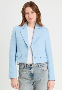 Veste courte bleu clair en tissu texturé, dotée d'un col cranté, de deux poches avant et d'une fermeture à boutons argentés, stylisée sur un top blanc.