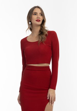 Červený crop top s dlouhými rukávy, vyrobený z lesklé tkaniny. V kombinaci se sukni se stehem do vysokého rozparku. Hladká textura.