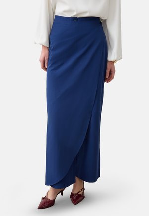 Mujer con falda larga azul de estilo cruzado con botones en la cintura, blusa blanca y zapatos de tacón altos puntiagudos color burdeos.