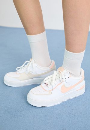 Osoba nosząca białe i jasnoróżowe sneakersy Nike Air z białymi skarpetkami stoi na jasnoniebieskim, wykładzinowym podłożu.