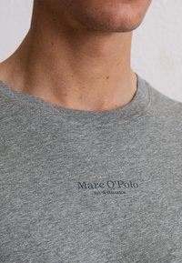 Zbliżenie na osobę w szarej koszulce Marc O'Polo, pokazujące dekolt oraz tekst marki na piersi.