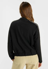 Person med långt brunt hår klädd i en svart randig jacka med vita axelstolpar och beige byxor, vänd bort från kameran mot en enfärgad bakgrund.