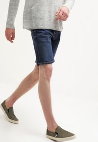 Seluar pendek denim berwarna biru gelap, potongan santai, dengan pinggiran yang digulung. Dipadankan dengan kasut slip-on zaitun dan atas lengan panjang berwarna kelabu terang.