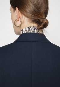 Námorně modrý blazer s texturovaným povrchem, s vzorovaným límcem v béžové a černé barvě. Zlatý náušnice ve tvaru kruhu viditelná na uchu modelky.