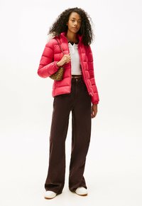 Czerwona kurtka puchowa z pikowanym wzorem, w połączeniu z białą bluzką i brązowymi, szerokimi spodniami. Modelka trzyma torbę w panterkę. Gładkie tło.