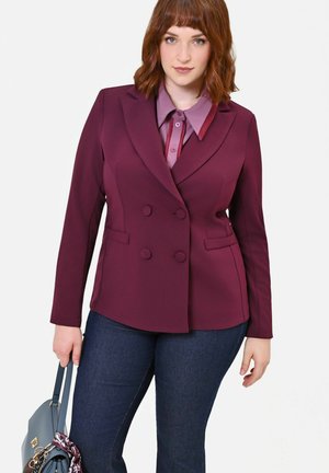 Donna che indossa una giacca doppiopetto bordeaux e una camicia viola, tiene una borsa blu, in piedi contro uno sfondo bianco.