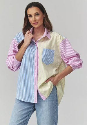 Kobieta o długich brązowych włosach, ubrana w oversize'ową, pasiastą koszulę w kolorach niebieskim, różowym i żółtym, z podwiniętymi rękawami i jasnoniebieskimi dżinsami, pozująca z ręką w kieszeni.