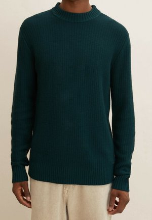 Pull en maille texturée vert foncé avec col rond côtelé, poignets et ourlet porté par une personne portant un pantalon beige clair.