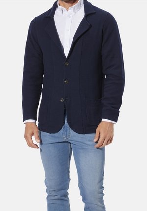 Hombre con blazer azul marino sobre una camisa blanca de botones y vaqueros azul claro, posando frente a un fondo blanco liso.