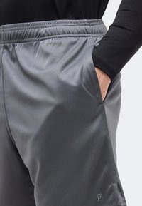 Άτομο που φοράει γκρι αθλητικό σορτς από δίχτυ με ελαστική μέση, χέρι στην δεξιά τσέπη, και μαύρο μακρυμάνικο τοπ ορατό.