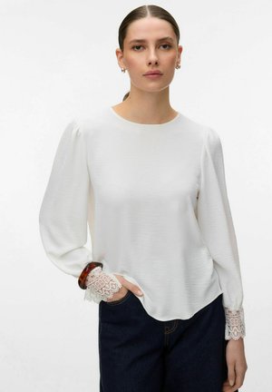 Femme aux cheveux plaqués en arrière, portant un chemisier blanc à manches longues avec des poignets en dentelle et un jean foncé, posant avec une main dans la poche.