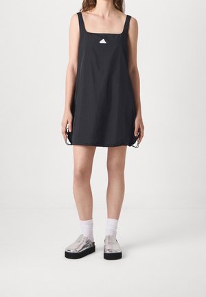 Kobieta w czarnej, bezrękawnikowej sukience z kwadratowym dekoltem, białych skarpetkach i metalicznych srebrnych butach na platformie typu slip-on, stojąca na jednolitym tle.