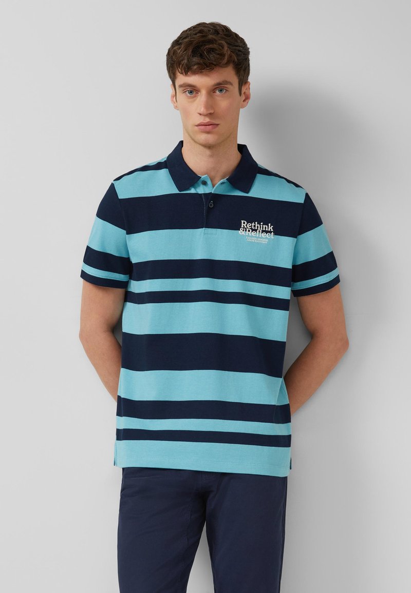 Jeune homme aux cheveux bruns courts portant un polo à rayures horizontales bleues et bleu marine avec le texte "Rethink & Reflect" sur la poitrine.