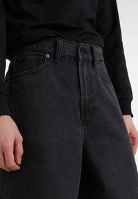 Jeans de mezclilla negra con un diseño de cintura alta y pierna recta, y una textura sutil. Incluye lazo para cinturón estándar y bolsillos frontales.
