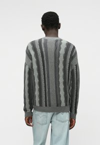 Γκρι πλεκτή ζακέτα με κάθετες ρίγες, υφασμάτινες λεπτομέρειες και ριμπ μανσέτες. Συνδυασμένη με ανοιχτό μπλε τζιν παντελόνι.