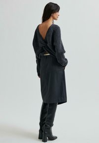 Žena v tmavém chlupatém svetru s otevřeným zkrouceným zádem, černé sukně ke kolenům a černých vysokých botách na podpatku stojící bokem.