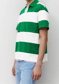 Henkilö, jolla on yllä vihreä- ja valkoraidallinen lyhythihainen polo-paita, yhdistettynä vaaleansiniset farkut, seisoo valkoista seinää vasten.