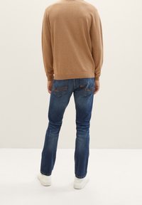 Suéter marrón de punto con dobladillo acanalado, combinado con jeans de mezclilla azules que presentan bolsillos traseros bordados y zapatillas blancas.