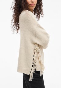 Κρέμα μαρινέ ζακέτα με φούντες στα πλαϊνά, μαλακή πλεκτή υφή, άνετη εφαρμογή και κοντά μανίκια. Συνδυασμένο με μαύρα παντελόνια.