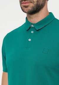 Homme avec barbe portant un polo vert avec deux boutons attachés et un petit logo carré cousu sur la poitrine gauche.