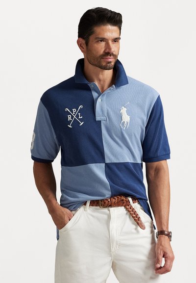 Mies, który ma na sobie niebieską koszulę polo w stylu color-block z wyszywanymi logo, białe spodnie, pleciony brązowy pasek i zegarek na nadgarstku, pozujący z rękami w kieszeniach.