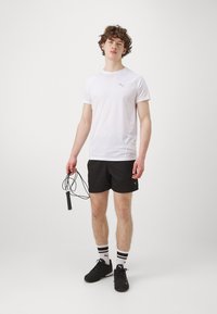 Valge lühikeste varrukatega sportlik särk halli logoga, mustad lühikesed püksid, mustad tossud ja triibulised sokid; hoiab hüpitsat, seistes ühtlasel taustal.