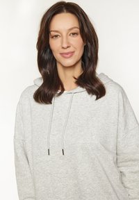 Hellgraue Kapuzenjacke mit großzügigem Design, Kordelzug am Hals und weicher Textur. Verfügt über eine lockere Passform und dezente Steppdetails.