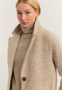Beige Strukturmantel mit weitem Kragen, einreihiger Knopfverschluss, kombiniert mit einem hellbeigen Rollkragenpullover mit Rippmuster.